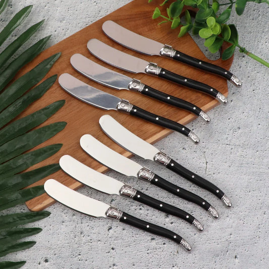 Set of six butter knives with black handles on a wooden board, surrounded by green leaves.